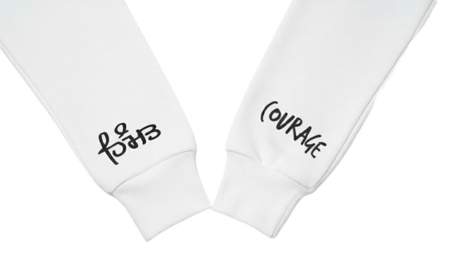 A close-up shot of the cuffs of two white hoodie sleeves. The left sleeve features black text in Gurmukhi "Himmat" translating to Courage. The right sleeve has the word "COURAGE" written in a stylized, handwritten black font. The cuffs are ribbed, and the background is plain white.