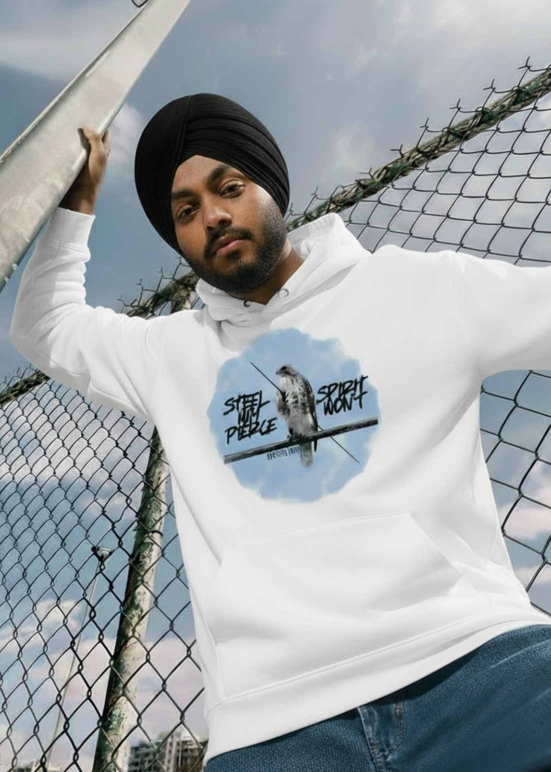A man in a black turban and white Cotton Fleece hoodie leaning against a chain-link fence under a blue sky. The hoodie features a circular blue watercolor-style graphic of a Baaz (Sikh falcon) perched on a branch, flanked by the edgy script "STEEL MAY PIERCE" and "SPIRIT WON'T.