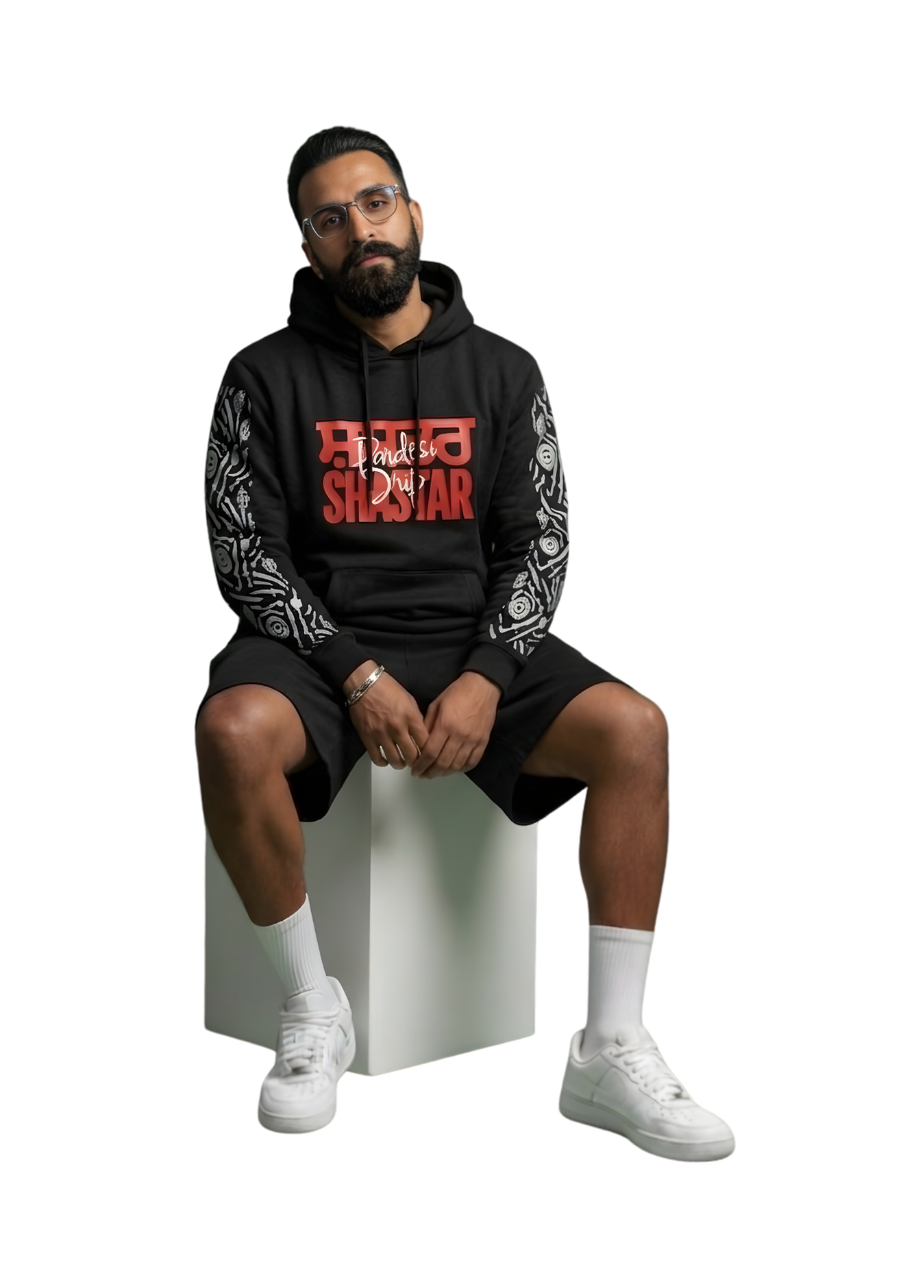 A portrait photograph of a South Asian man wearing a black "SHASTAR" graphic hoodie, a silver bracelet, black shorts, white socks, and white sneakers, sitting on a white block against a green studio background.