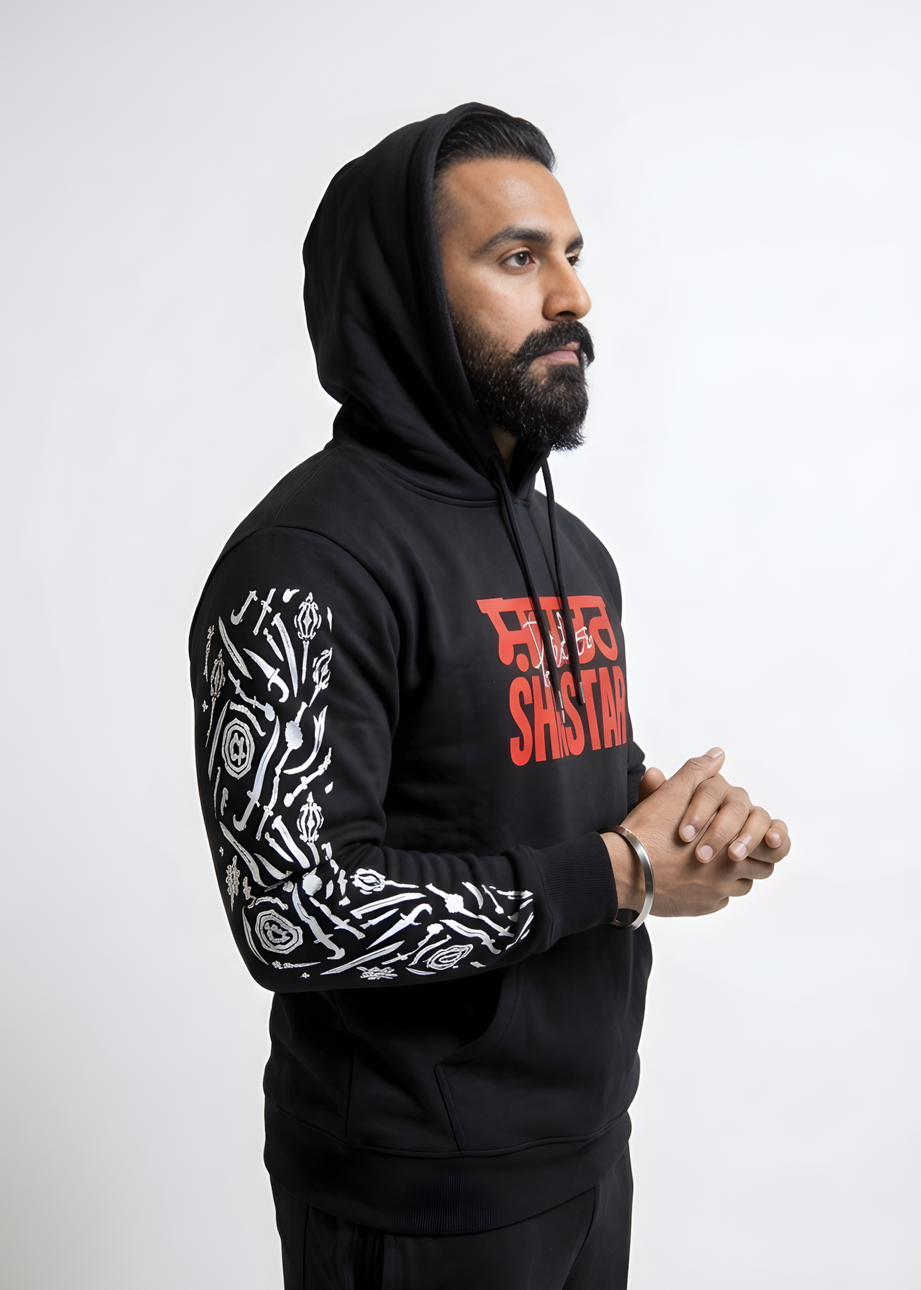 Black Punjabi streetwear hoodie featuring "Shastar" graphic print in red and white Shastar sleeve art. Model with beard and silver Kara wearing a hooded sweatshirt with Gurmukhi calligraphy and Sikh warrior weapon designs.
