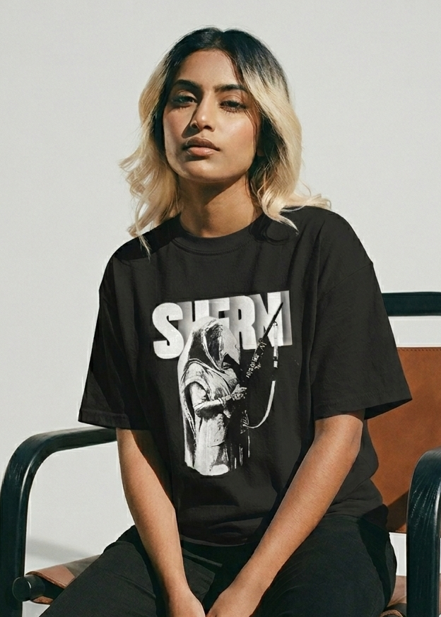 South Asian female wearing a black oversized French terry T-shirt with a graphic design sitting on a chair against a plain background.