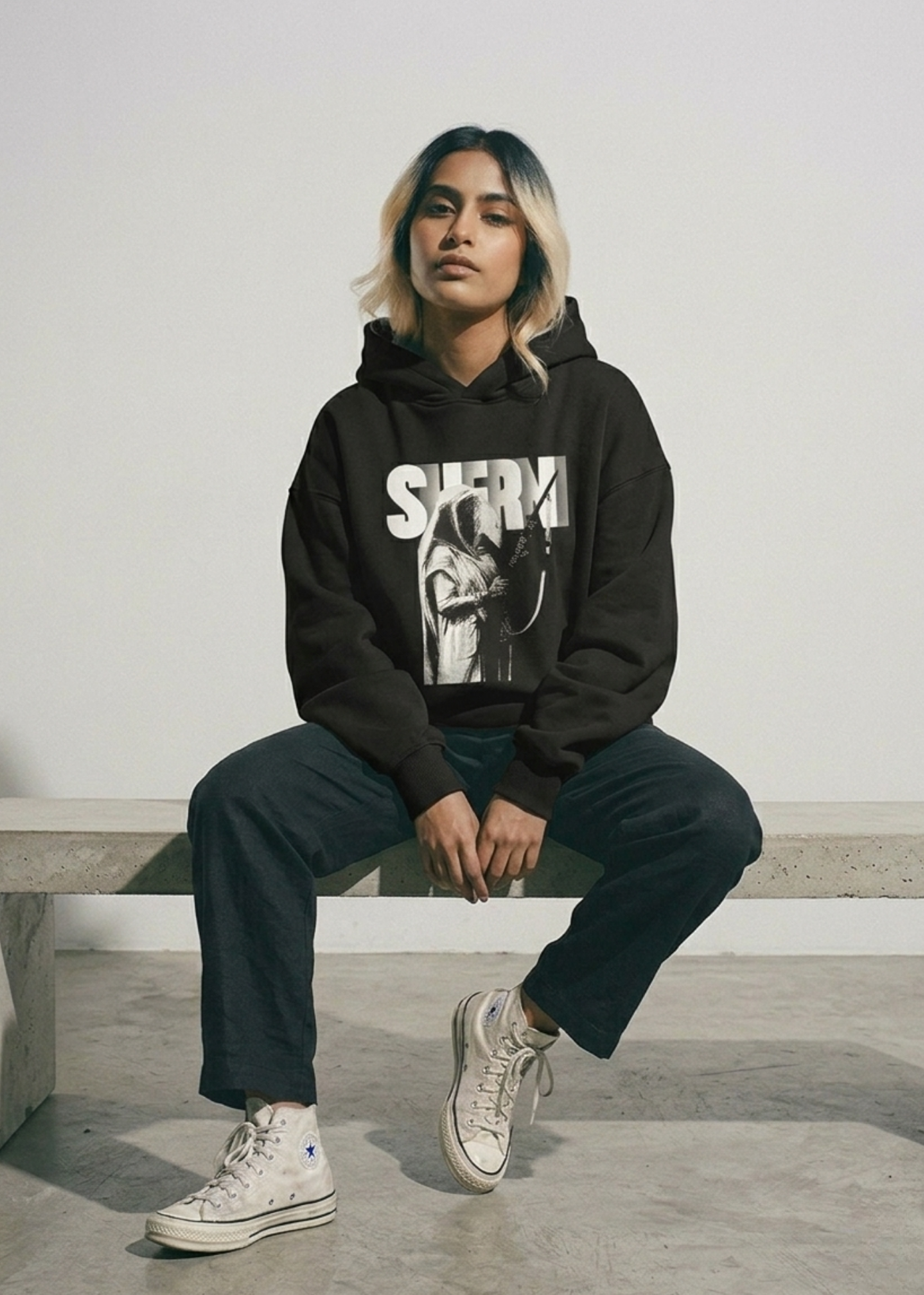 A woman with blonde-streaked hair sitting on a concrete bench, wearing a black oversized Cotton Fleece hoodie and high-top sneakers. The hoodie features a large, bold graphic of a historical figure, Maharani Jindan Kaur, with the word "SHERNI" in large white block typography in the background. The design is loud, graphic, and culturally inspire.