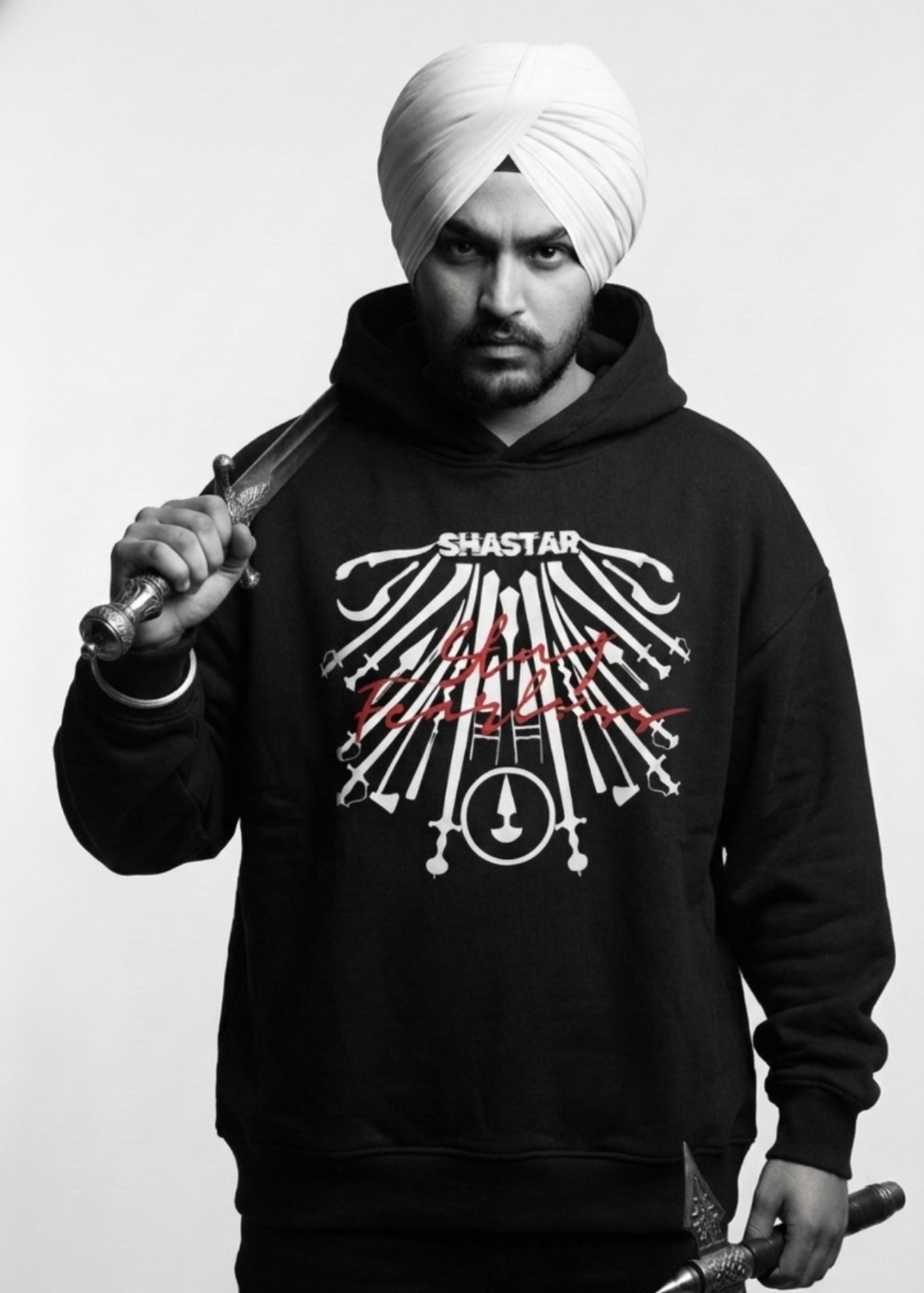 Sikh turbaned male wearing a black Cotton Fleece Hoodie with a white graphic design featuring panjabi Shastar with "Stay Fearless" written in red on a plain background.
